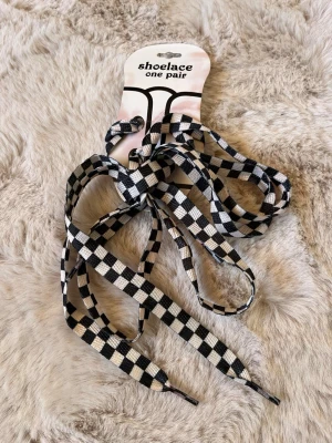 Schackrutiga skosnören svart/vit - Ett par platta skosnören med svart och vitt schackrutigt mönster. Perfekt för att ge dina sneakers eller kängor en unik och personlig touch. Tillverkade i slitstarkt tygmaterial och passar de flesta skotyper.