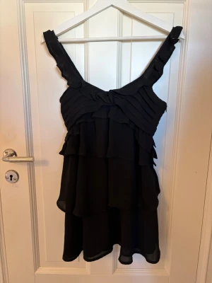 Svart volangklänning med breda axelband - Säljer en svart kortklänning med breda axelband och flera lager volanger. Klänningen har en omlottliknande överdel och är tillverkad i ett lätt, flygigt material som ger en snygg siluett. Perfekt för dig som gillar en feminin och trendig stil.