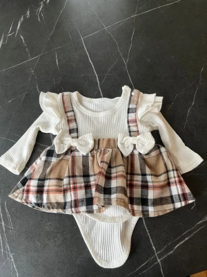 Rutigt bodyset med volanger från SHEIN - Supersött bodyset från SHEIN med långärmad ribbad vit topp, volangdetaljer vid axlarna och fastsydd kjol i beige, svart och röd rutigt mönster. Två dekorativa rosetter framtill och praktiska knappar i grenen. Perfekt för en gullig look!