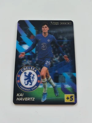 Kai Havertz Premier League fotbollsskort   - Samlarkort från Panini med Chelsea FC-logga, visar en spelare i blå matchtröja och klubbens logga. Kortet har färgstarka blå och lila detaljer samt texten 'Kai Havertz' och '+5' i hörnet. Perfekt för dig som gillar fotboll och att samla på kort. Pris kan diskuteras! 