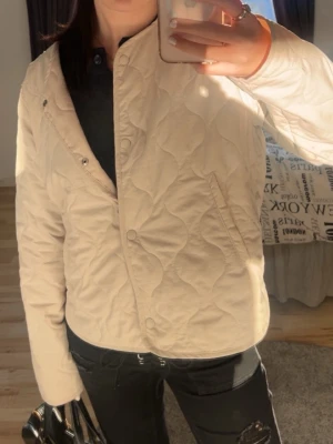 Beige quiltad cardiganjacka - Snygg beige quiltad cardiganjacka med tryckknappar och diskret mönster. Jackan har två fickor framtill och en rak passform. Perfekt för lager-på-lager och ger en clean look till din outfit.