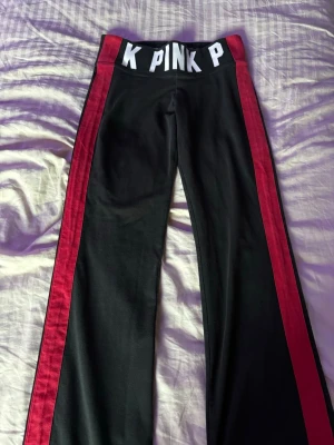 Svarta mjukisbyxor från VS PINK - Snygga svarta mjukisbyxor från VS PINK med röda ränder längs sidorna och bred resår i midjan med vit text. Byxorna är lite bootcut och är perfekta för en avslappnad stil. Materialet känns mjukt och bekvämt. Byxorna är i storlek S men funkar jätte bra på XS också! 