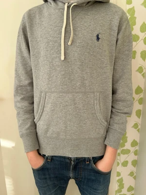 Grå hoodie från Polo Ralph Lauren - Snygg grå hoodie från Polo Ralph Lauren med klassisk marinblå logga på bröstet. Hoodien har huva med vita dragsnören och en stor magficka. Tillverkad i mjukt bomullsmaterial som känns skönt mot huden. Perfekt till jeans för en avslappnad look. Nypris ca 2000 kr