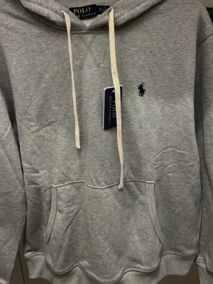 Grå hoodie från Polo Ralph Lauren - Snygg grå hoodie från Polo Ralph Lauren med klassisk huva och det ikoniska mörkblå polospelar-loggan broderad på bröstet. Tillverkad i mjukt bomullsmaterial som är perfekt för chill dagar. Enkel och stilren design som funkar till det mesta. Den är lite stor k storleken