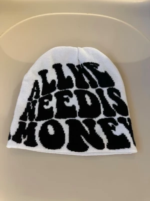 Vit mössa med svart textprint - Säljer en vit mössa med stor svart text som säger 'ALL WE NEED IS MONEY' i fet, grafisk stil. Mössan är stickad och har en avslappnad passform. Perfekt accessoar för dig som gillar statement-plagg och streetwear vibes.
