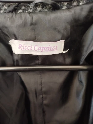 Grå rutig kappa från Ricci Capricci - Stilren grå kappa från Ricci Capricci med klassiskt rutmönster. Kappan har en rak passform, krage och är tillverkad i ett tjockare tyg som passar perfekt för kyligare dagar. Snygg att matcha med både jeans och boots.200kr