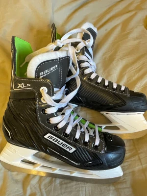 Bauer XLS - Säljer ett par svarta Bauer XLS skridskor med vita snören och detaljer i vitt och grönt. Skridskorna har en robust plastkonstruktion och klassisk design med hög komfort och stöd runt ankeln. Perfekta för hockey eller fritidsåkning på is.
