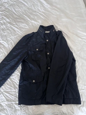 Mörkblå vindjacka från Nils Sundström - Snygg mörkblå vindjacka från Nils Sundström, silverfärgade knappar och dragkedja. Jackan har flera fickor framtill för en cool look. Perfekt för dig som vill ha en stilren och funktionell jacka. Tunn och skön för våren!