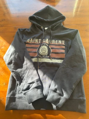 Mörkblå Saint Laurent hoodie - Snygg blå hoodie från Saint Laurent med tryck i vitt och rött framtill. Klassisk huva med dragsko och magficka. Trycket har en vintage-look med texten SAINT LAURENT och SL-emblem. Perfekt för dig som gillar streetwear och vill sticka ut.