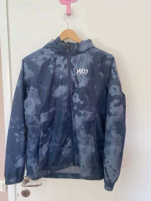 Mörkblå vindjacka från WARP - Snygg mörkblå vindjacka från WARP med vattenliknande mönster och hel dragkedja framtill. Jackan har huva och två sidofickor, perfekt för blåsiga dagar. Materialet är tunt och lätt, vilket gör den enkel att bära med sig. Logga på bröstet i vitt.