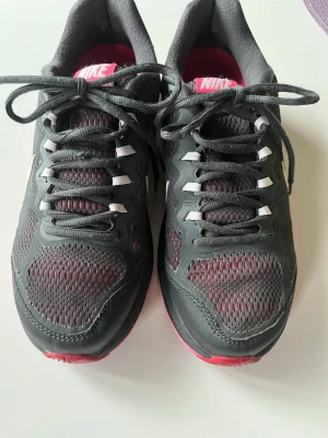 Nike grå sneakers med rosa detaljer - Snygga sneakers från Nike i mörkgrått med meshpaneler för bra ventilation och rosa detaljer på insidan och sulan. Skorna har klassisk snörning, vit Nike-logga på hälen och en sportig look. Perfekta för dig som gillar streetstyle och komfort.