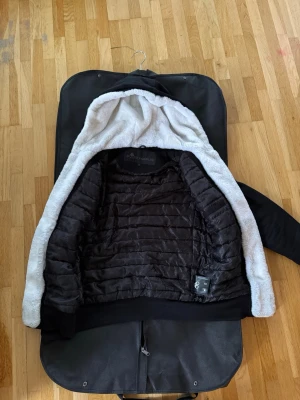 Svart teddyjacka med huva från Moose Knuckles - Svart teddyjacka från Moose Knuckles med vit fluffig foder och stor huva. Jackan har dragkedja framtill, snörning i huvan och fickor på magen. Ärmarna är långa och jackan har ribbade muddar. Perfekt för kyliga dagar.