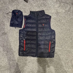 Mörkblå dunväst från Polo Ralph Lauren - Snygg mörkblå dunväst från Polo Ralph Lauren med röd insida och röda detaljer på dragkedjorna. Västen har broderad röd logga på bröstet och medföljer en matchande påse. Perfekt för lager-på-lager och chill stil.