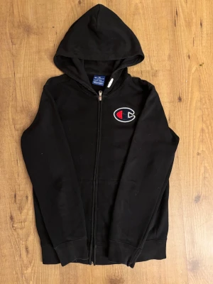 Svart hoodie från Champion med dragkedja barnstorlek - Säljer en svart hoodie från Champion med dragkedja framtill och stor logga på bröstet. Hoodien har huva och två fickor framtill. Tillverkad i mjukt bomullsmaterial som är skönt att ha på sig. Perfekt för en avslappnad och sportig stil. Passar nån runt 150/155 cm lång