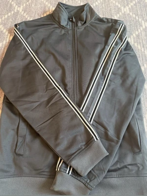 Grå track jacket med vita ränder - Snygg grå track jacket med vita och svarta ränder längs ärmarna. Jackan har hög krage, dragkedja framtill och ribbade muddar vid ärmslut och nederkant. Perfekt för en sportig och avslappnad stil.