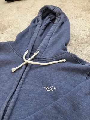 Blå zip hoodie från Hollister - Mysig blå hoodie från Hollister med dragkedja och vit broderad logga på bröstet. Tröjan har huva med vita snören och är gjord i mjukt sweatshirtmaterial. Perfekt för chill dagar eller när du vill ha en avslappnad look.