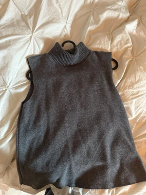 Grå stickad poloväst  - Mysig grå stickad väst med hög polokrage. Västen är ärmlös och har en rak, loose passform som passar perfekt över en skjorta eller t-shirt. Enkel och stilren design utan detaljer eller mönster. (PRIS KAN DISKUTERAS