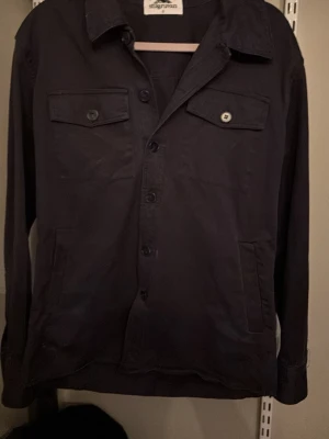 Svart overshirt från Stilgruvan - Stilren svart overshirt från Stilgruvan med klassisk krage, knäppning framtill och två bröstfickor med lock och knapp. Tillverkad i ett kraftigare bomullstyg som ger en schysst och avslappnad look. Perfekt att bära över en t-shirt eller hoodie.