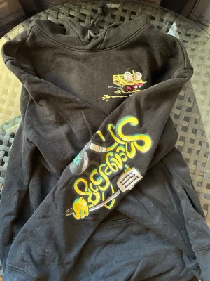 Svart hoodie med Spongebob-tryck - Svart hoodie med stor färgglad Spongebob-grafik på ryggen och texten 'Stay Pretty' i lila och gult. Tryck även på bröstet och ena ärmen med tecknad stil och coola detaljer. Huva med dragsko och känguruficka framtill. Perfekt för dig som gillar streetwear och unika prints.