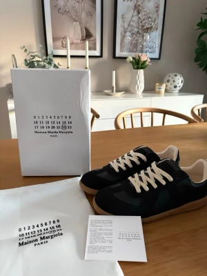 Maison Margiela svarta sneakers - Svarta sneakers från Maison Margiela med beigea skosnören och klassisk gummisula. Skorna har en låg silhuett och är tillverkade i en mix av mocka och läder. Snygg minimalistisk design med diskreta detaljer och ikonisk Margiela-märkning.