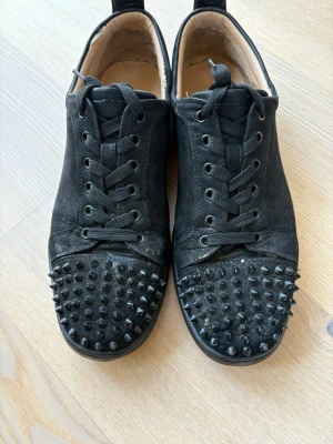 Louboutin - Coola svarta sneakers från Christian Louboutin med nitar på tån och klassisk snörning. Skorna har en låg profil och är tillverkade i textil med läderdetaljer på insidan. Perfekt för dig som vill sticka ut med en edgy stil.