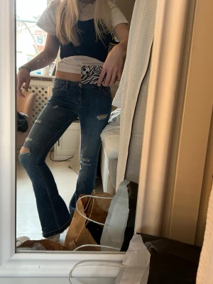 Blå slitna bootcut jeans - Säljer ett par blå bootcut jeans med slitningar och hål på knäna. Jeansen har låg midja och klassisk femficksdesign. Perfekta för en avslappnad och trendig look. Materialet är denim i bomull.