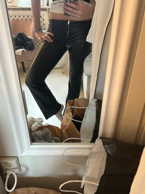 Svarta skinnbyxor med dragkedja - Säljer ett par svarta skinnbyxor med rak passform och hög midja. Jeansen har en synlig dragkedja framtill och sitter tight upptill men är lite vidare nedtill. Materialet är glansigt och ger en cool look.
