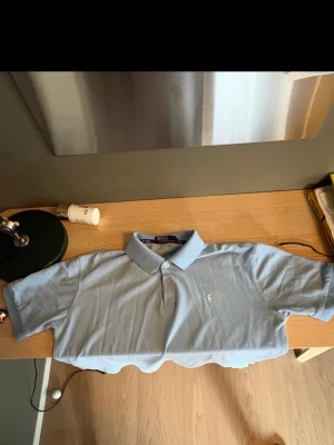 Ljusblå piké från Ralph Lauren - Klassisk ljusblå pikétröja från Ralph Lauren med korta ärmar och vit broderad logga på bröstet. Tröjan har krage och knappar framtill. Perfekt för en clean och stilren look. Storlek M