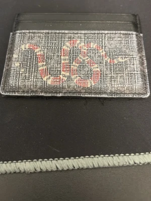 Gucci korthållare med orm-motiv - Snygg korthållare från Gucci i grått canvasmaterial med det klassiska ormmotivet i rött, vitt och svart på framsidan. Plats för flera kort och stilren design med exklusiv känsla. Perfekt accessoar för dig som gillar lyxiga detaljer.