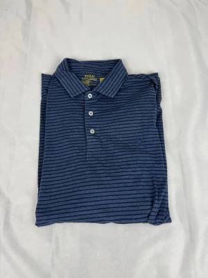 Blå randig piké från Polo Ralph Lauren - Snygg blå pikétröja från Polo Ralph Lauren med tunna mörkblå ränder och klassisk krage. Tröjan har tre knappar framtill och är tillverkad i ett mjukt bomullsmaterial. Perfekt för en avslappnad men stilren look.