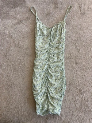 Blommig kortklänning med smala band - Söt kortklänning med smala axelband och rynkade detaljer framtill. Klänningen har ett ljust blommigt mönster i grönt och vitt samt en liten knytning vid bysten. Perfekt för varma dagar och sommarens alla tillfällen.