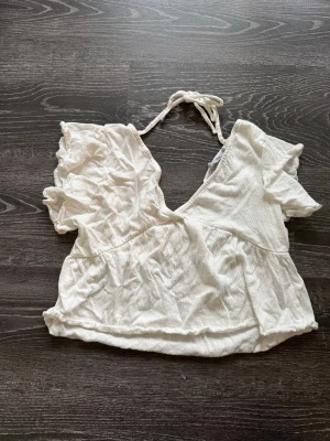 Vit omlottopp med knyt och volang - Söt vit omlottopp med korta volangärmar och knyt i nacken. Toppen har en djup v-ringning och är croppad, vilket ger en trendig och luftig känsla. Perfekt för varma dagar och enkel att styla med jeans eller kjol.