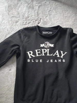 Svart sweatshirt från Replay - Cool svart sweatshirt från Replay med vit logga och texten 'Replay Blue Jeans' på bröstet. Tröjan har rund halsringning och är tillverkad i mjukt material som känns skönt mot huden. Perfekt för en avslappnad och trendig look.