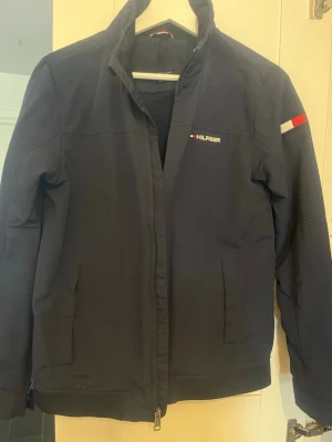 Marinblå vindjacka från Tommy Hilfiger - Snygg marinblå vindjacka från Tommy Hilfiger med diskret logga på bröstet och ärmen samt tryckt  text på kragen. Jackan har dragkedja framtill, två sidofickor och en klassisk ståkrage. Perfekt för dig som vill ha en stilren och sportig look.