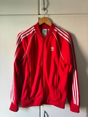 Röd Adidas track jacket - Klassisk röd Adidas track jacket med vita ränder längs ärmarna och vit logga på bröstet. Dragkedja framtill, ribbade muddar och krage. Tillverkad i polyester för sportig känsla och retrostil. Köpte detta addidas sättet för 2 år sedan, tyvärr gick byxorna sönder och tröjan håller nu på att bli för lite för mig, har därför valt att sälja den. 