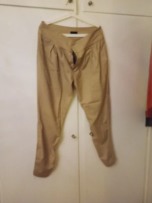 Beige chinos med knappar framtill - Snygga beige chinos med avslappnad passform och knäppning framtill. Byxorna har veckade detaljer vid midjan och är tillverkade i ett lätt bomullsmaterial. Perfekta för en casual och trendig look.