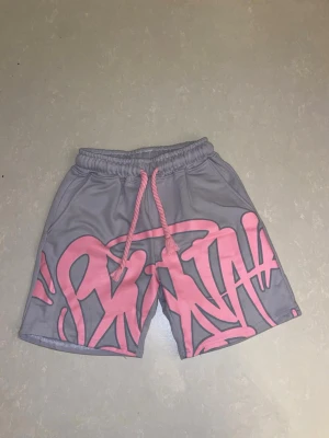 Syna World shorts - Säljer ett par grå shorts med fett rosa graffiti-tryck över hela framsidan. De har elastisk midja med rosa dragsko och sidofickor. Perfekta för chill dagar eller streetwear-stil. Materialet känns mjukt och lätt, troligen polyester eller bomullsblandning.