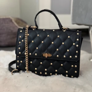 Väska med nitar  - Snygg svart handväska i quiltat konstläder med guldfärgade nitar och kedjedetalj. Väskan har ett top handtag och en axelrem i guld, samt ett vridlås framtill. Perfekt accessoar för att lyfta din outfit.