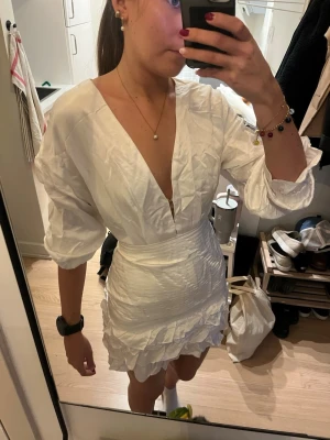 Vit klänning med volang - Säljer en vit kortklänning med djup v-ringning. Klänningen har långa ärmar och volangdetaljer längs nederkanten. Tillverkad i ett silke material som ger en lyxig känsla. Perfekt för dig som vill sticka ut med en elegant och trendig look. Den behöver strykas!