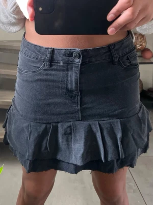 Mörkgrå jeans volangkjol - Säljer en mörk grå kortkjol med volangdetaljer och jeanslook. Kjolen har klassiska fickor fram och bak, bälteshällor och stängs med knapp och dragkedja. Perfekt för dig som gillar en edgy och trendig stil.