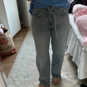 Grå raka jeans med knytskärp - Säljer ett par grå jeans med svaga barrel ben och hög midja. Jeansen har ett matchande knytskärp i midjan. Aldrig använda och lappen sitter kvar