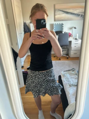 Svart blommig kortkjol med volang - Supersöt svart kortkjol med vitt blommönster och volangkant. Kjolen har en flowy passform och är perfekt för varma dagar. Tyget känns lätt och skönt mot huden. Kjolen har en dragkedja på sidan (syns på sista bilden) vilket gör den enkel och bekväm att ta på och ta av. Kjolen har även en resor på insidan av midjan vilket gör den lite extra stretchig💕 passar en xxs-xs och säljer då jag har väldigt många kjolar 