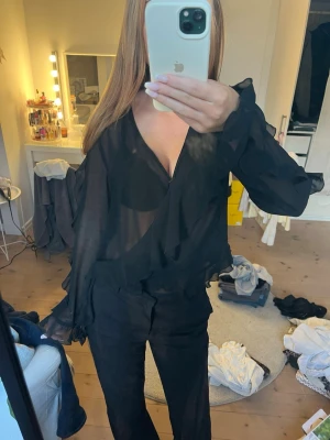 Svart transparent blus med volanger - Säljer en svart, transparent blus med v-ringning och volangdetaljer längs halsringningen. Blusen har långa ärmar med lätt utsvängda ärmslut och är gjord i ett tunt, luftig material som ger en snygg och trendig look. Har satt en säkerhets nål vid bröstet men är bara att ta bort, den blir lite för urringad annars!