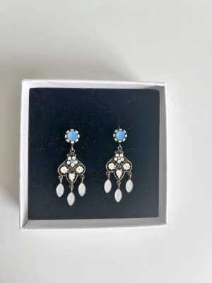 Blå och vita pärlörhängen - Unika, anrika örhängen, dekorerade med ljusblå stenar och vita pärlor. Hängande design med tre droppformade pärlor på varje örhänge och detaljer i silverfärgad metall. Perfekta för dig som gillar vintageinspirerade accessoarer.