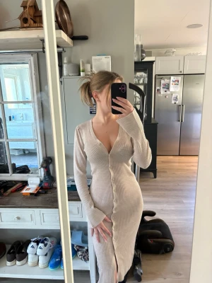 Beige ribbad långklänning med knappar - Säljer en beige ribbad långklänning med v-ringning och långa ärmar. Klänningen har knappar hela vägen framtill och en liten slits nedtill. Perfekt för dig som gillar en stilren och enkel look. Obs på mig är den ankel lång o jag är 165 cm lång, stl Xs