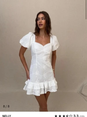Studentklänning från Nelly  - Superfin vit kort klänning från Nelly med broderade blommor, puffärmar och volangkant nertill. Klänningen har öppen rygg med stor rosett och söta spetsdetaljer vid halsringningen. Perfekt till studenten!