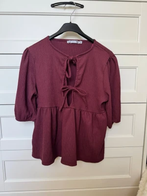 Vinröd blus med knytning fram - Vinröd blus från Lager 157 med trekvartsärmar och två knytband framtill. Blusen har en lätt rynkad struktur och en lös passform med markerad midja. Materialet känns mjukt och luftigt, perfekt för att skapa en avslappnad look.