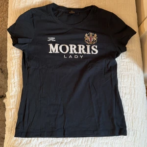 Svart Morris Lady t-shirt - Snygg svart t-shirt från Morris Lady med broderad logga och emblem på bröstet. Klassisk passform och rund halsringning. Tillverkad i mjuk bomull som känns skön mot huden. Perfekt för en stilren och enkel look.