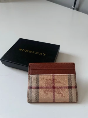 Burberry rutigt kortfodral i läder - Säljer ett stilrent kortfodral från Burberry i klassiskt beige, brunt och rött rutmönster. Tillverkat i läder med plats för flera kort. Perfekt accessoar för dig som gillar lyxiga detaljer och ikonisk design. Kommer med original ask.