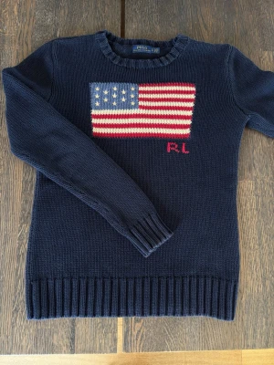 Mörkblå stickad tröja Polo Ralph Lauren S - Säljer en mörkblå stickad tröja från Polo Ralph Lauren med amerikansk flagga i rött, vitt och blått på bröstet samt RL-broderi. Tröjan har rund halsringning och ribbade muddar vid ärmslut och nederkant. Perfekt för dig som gillar klassisk preppy stil. Storlek S